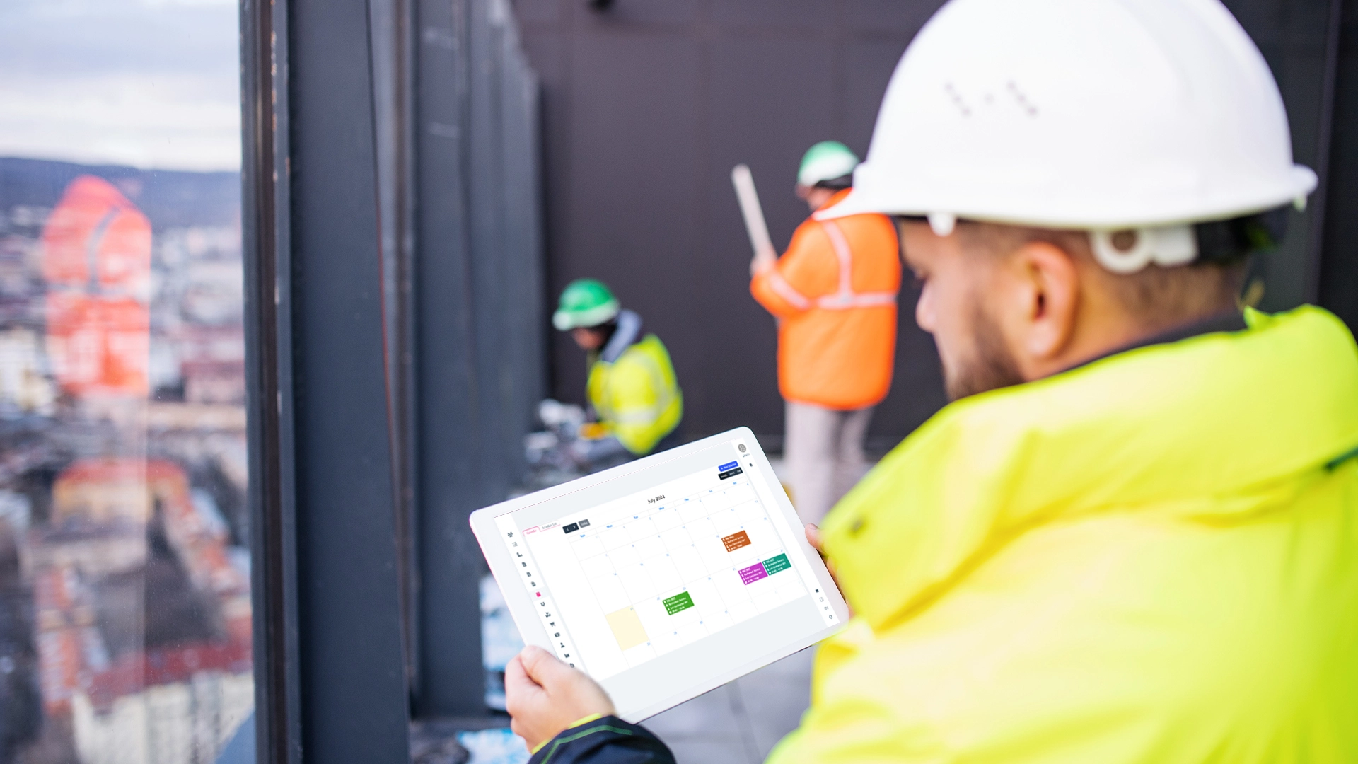 A construction worker in a hard hat and high-visibility jacket using a tablet with scheduling screen of homearize flooring software , while other workers in safety gear operate in the background on a construction site.