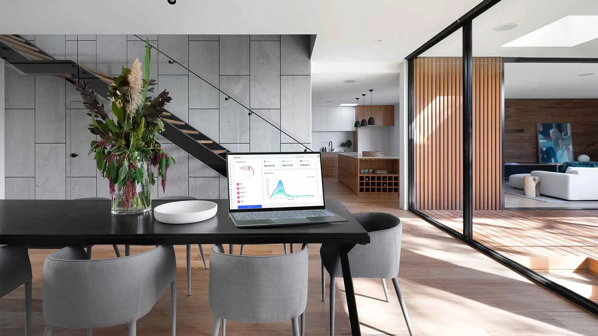 A modern home interior with a dining table featuring a laptop displaying a homearize flooring software dashboard, surrounded by elegant gray chairs and a large vase of flowers, with a wooden kitchen and living area in the background.